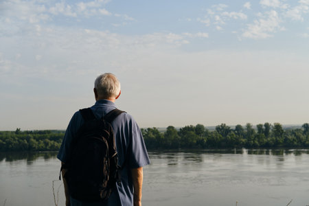 Elderly gray-haired man stands on observation deck with tourist backpack and admires the Danube River, in old town Zemun at dawn. Rear view. Serbia, Belgrade - 04.16.2024.の写真素材