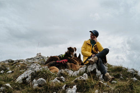 Travel and hiking with pet concept. Young woman sits on hill with German and Australian shepherd dogs. Traveler in a yellow raincoat and admires view of mountains. Montenegro, Durmitor national parkの写真素材