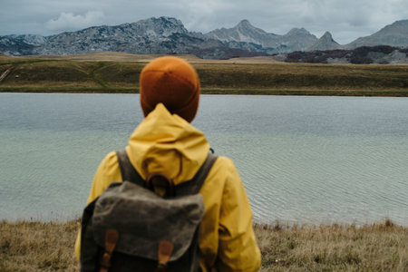 A young traveler woman wearing yellow raincoat and retro style backpack, hiking in mountains and...