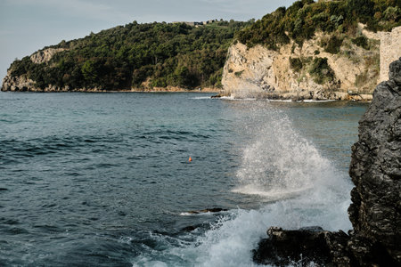 Dramatic coastal scene where a wave crashes against jagged rocks. The deep blue sea stretches towards a lush, green hillside, with rugged cliffs adding depth and texture to the landscape.の写真素材