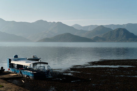 Morning light over Skadar Lake in Montenegro with a moored blue boat and calm water surrounded by misty mountain silhouettes. Peaceful and scenic lakeside atmosphereの写真素材