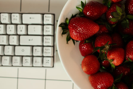 Overhead close-up of a white bowl brimming with ripe red strawberries beside a compact white...