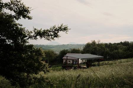 Pastoral countryside view with green hills, trees, and rooftops under cloudy skies. Serbian village in spring season. Small farmhouse with tin roof among grassy hills and distant mountains at sunsetの写真素材