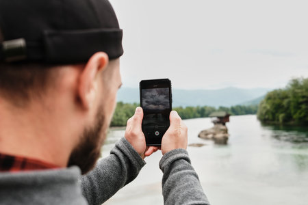 Close-up of a man filming the Drina River house on his phone, capturing the iconic Serbian view. Rear view. A popular tourist destination. Serbia country in spring seasonの写真素材