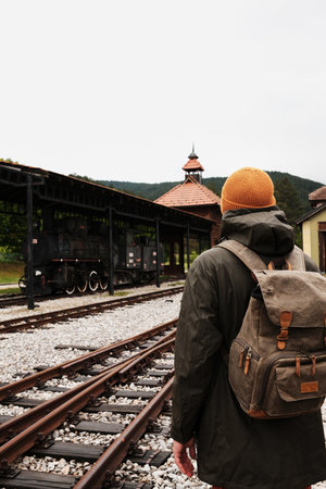 Male traveler in yellow hat with backpack walking near train tracks at Mokra Gora station, Serbia....