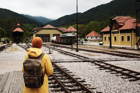Woman in yellow jacket and orange hat at Mokra Gora railway station, looking at mountain landscape. Rear view. Solo travel concept.の写真素材