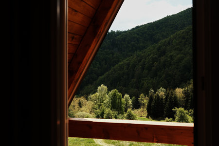 Looking through a triangular attic window toward green wooded hills. A-frame cabin in Tara National Park, Serbia. Peaceful nature and architectural charm combined.の写真素材