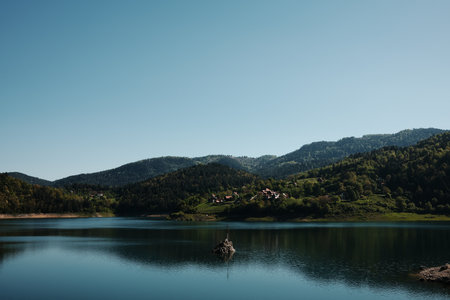 Calm waters of Zaovine Lake surrounded by forested hills in Tara National Park during spring. Serbia country. There is a large iron cross set in stone in the middle of the lakeの写真素材