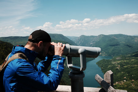 Man in blue jacket looking through a telescope at the lush hills and winding Drina River in Tara National Park, Serbia. Binoculars for tourists on viewpointの写真素材