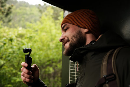 A man in a beanie and jacket films the forest landscape with a small camera from a moving train in...