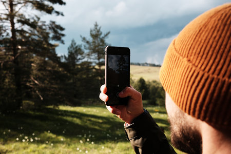 A man in an orange beanie holds a smartphone to take a photo of the scenic forest and meadow...