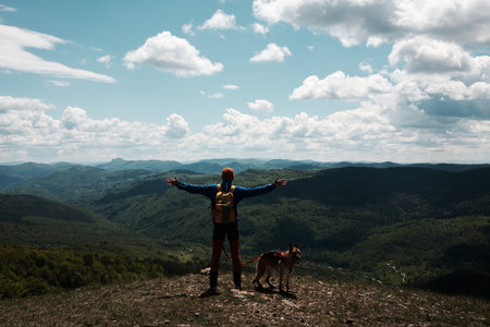 Man with backpack standing with arms open beside his dog on Mount Cigota, enjoying freedom and fresh mountain air. Traveler with German shepherd. Rear view. Travel with pet conceptの写真素材