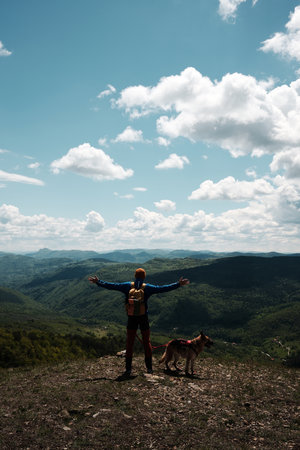 Man with backpack standing with arms open beside his dog on Mount Cigota, enjoying freedom and fresh mountain air. Traveler with German shepherd. Rear view. Travel with pet conceptの写真素材