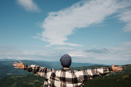 A woman stands with arms open, enjoying a panoramic mountain view under a wide spring sky. Rear view