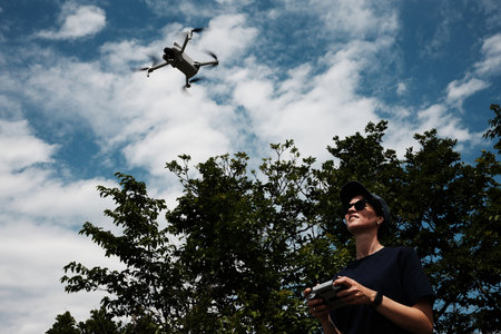 Woman in sunglasses pilots a drone under the blue sky. She stands near green trees with a focused...