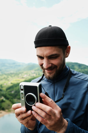 Man smiling while using an instant retro camera outdoors, with forested mountains in the background