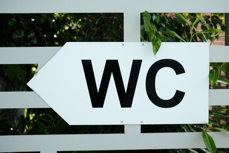 White directional sign with black WC letters pointing left, mounted on a fence with green leaves in...