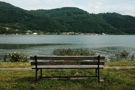 A wooden bench facing a river and green hills, surrounded by wildflowers and grassの写真素材