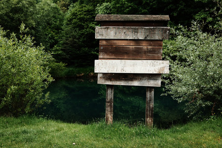 Weathered wooden sign on stilts stands by a calm, green river surrounded by forest. A scene from a tranquil nature reserve. Copy space for travel and outdoor designsの写真素材