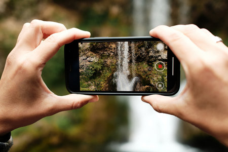 Hands holding smartphone filming Veliki Buk waterfall in Lisine reserve, Serbia. Natural wonder...