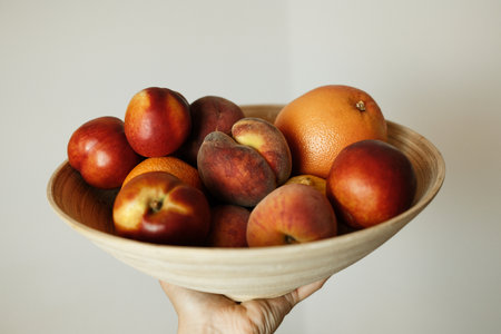 Hand holding a wooden bowl full of fresh summer fruits - nectarines, peaches, oranges and grapefruit against a neutral indoor background.の写真素材