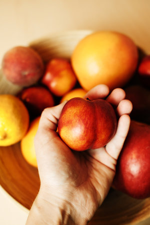 Hand holding a ripe nectarine over a wooden bowl filled with mixed fruits including grapefruit, lemon, and peaches. Healthy lifestyle and nutrition conceptの写真素材