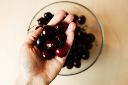 Close-up of a hand holding a handful of fresh cherries over a glass bowl. Juicy summer fruit ready to eatの写真素材