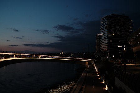 Night view of the Belgrade Waterfront promenade with modern high-rises, bike path, and dramatic sky....