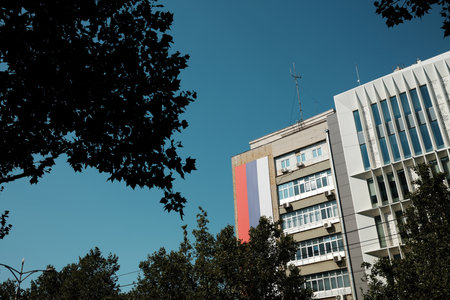 Modern office building in Belgrade with a flag on the wall, clear blue sky and trees in the...