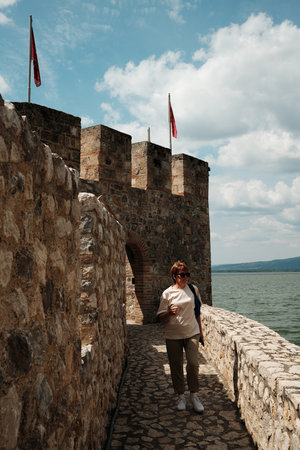 Tourist woman walks along the stone path at Golubac Fortress with flags flying above the medieval walls. Travel in Serbia conceptの写真素材
