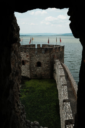 Scenic view through Golubac castle window of a round stone tower with flags above a green moat and wide river. Peaceful historical architecture. Serbia countryの写真素材