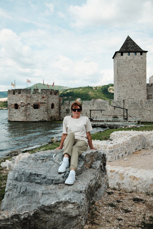 Woman sitting on a large rock with Golubac Fortress in the background. Medieval stone walls and Danube River view. Travel in Serbia conceptの写真素材