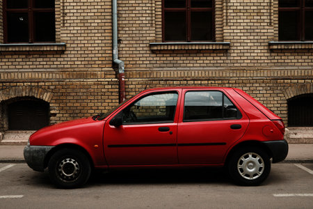 Old red compact car parked in front of a brick building wall in Sombor, Serbia. Urban street scene