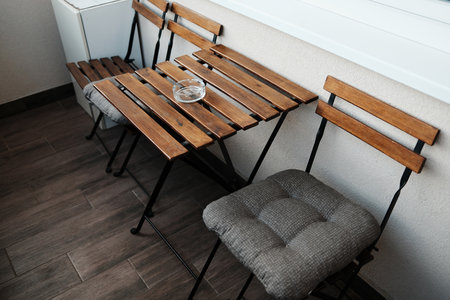 Small balcony with wooden chairs and table set, featuring a glass ashtray and cozy cushion, shot in natural lightの写真素材