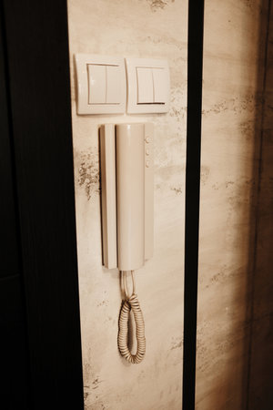 Wall-mounted intercom telephone with coiled cord next to two light switches on a textured wall in...