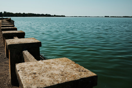 Stone fence and calm water of Lake Palic, Serbia, under blue sky. Tranquil landscape and peaceful...