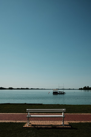 A white bench faces Lake Palic under a clear blue sky in Serbia. Calm water, minimalism, and peaceful scenery with a floating dock in the distanceの写真素材
