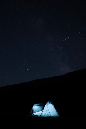 Illuminated camping tent under a starry night sky with visible meteors, creating a peaceful outdoor...