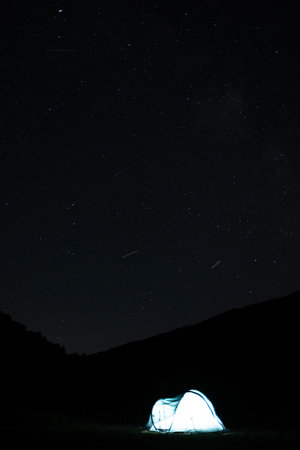 Illuminated camping tent under a starry night sky with visible meteors, creating a peaceful outdoor astrophotography scene. Outdoor travel concept. View of the Milky Way from the northern hemisphereの写真素材