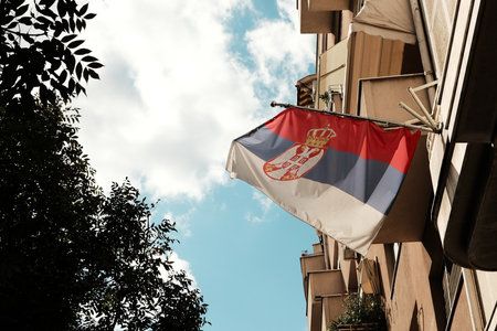 Serbian national flag with crown waving from a residential building against blue sky and trees. Patriotic symbol in everyday urban lifeの写真素材