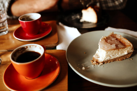 Slice of cheesecake on a plate with two cups of black coffee in red and orange mugs on wooden table. Cozy date in cafe sceneの写真素材