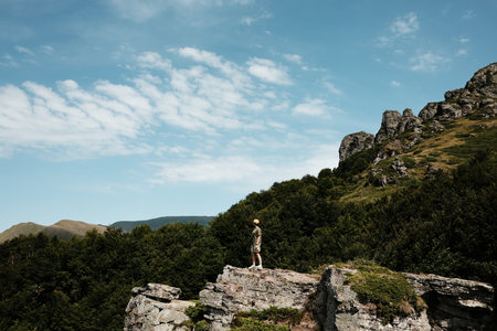 A young woman stands on a cliff in Stara Planina, Serbia, admiring vast green mountains under a blue sky. A perfect moment of freedom and adventure in natureの写真素材