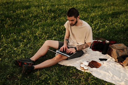 Man sitting on a blanket in the park working on a laptop, surrounded by tea set and backpack. Digital nomad conceptの写真素材
