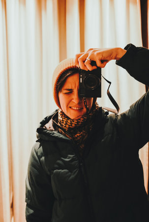 A young woman in a warm jacket and beanie takes a close-up mirror selfie with a retro film camera,...