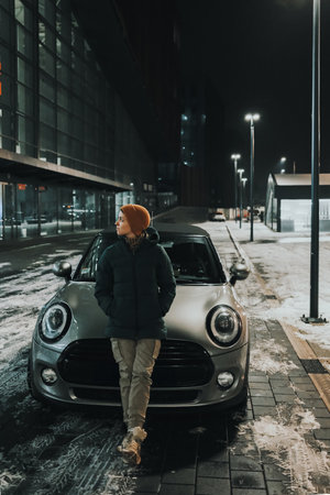 Woman in orange beanie leaning on silver car at night in winter snowy city. Female driver rests...