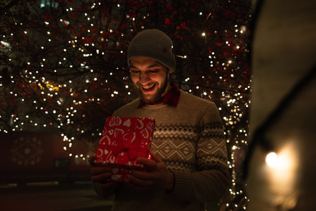 A smiling man in a knitted sweater and beanie opens a red Christmas gift box under festive holiday...