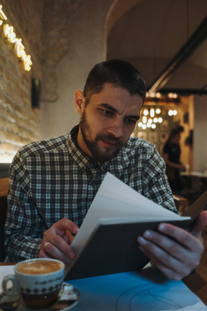 Man in a cozy cafe reading a menu with a cup of coffee on the table. Warm lights and rustic brick wall create a relaxed moodの写真素材
