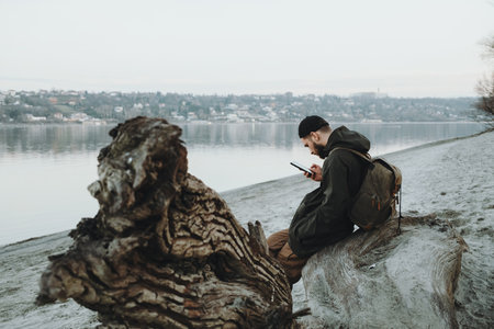 Young man with backpack sitting on driftwood at the Danube Riverbank in Novi Sad, using his phone during a cold winter dayの写真素材