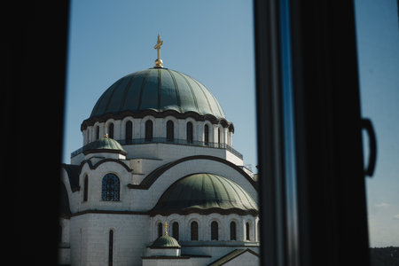 View of Saint Sava Temple in Belgrade framed by a window, with its green domes and golden cross shining under the blue skyの写真素材