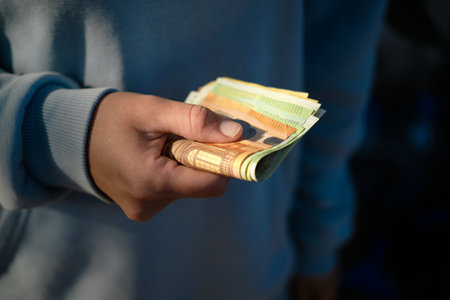 Person counting euro banknotes, holding 100 euro bill in focus with 50 and 200 euro notes in the backgroundの写真素材